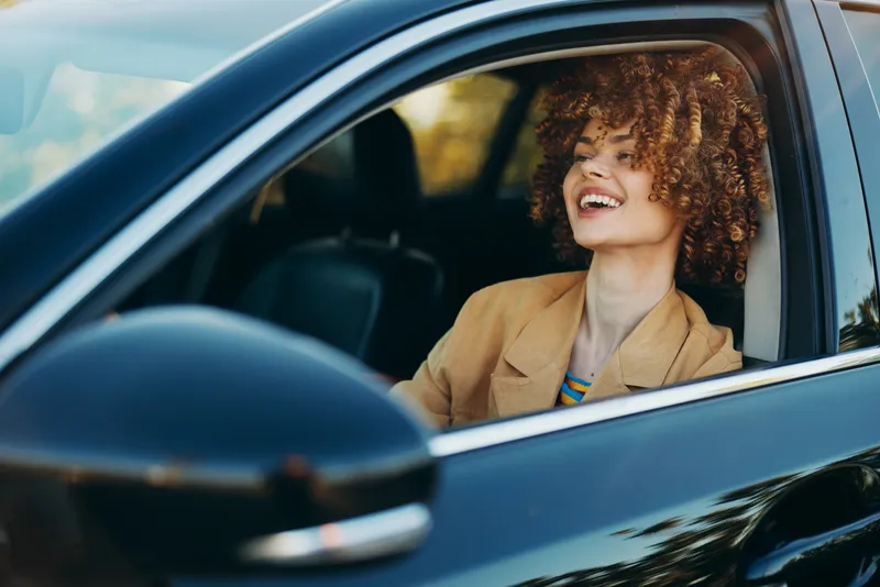 Young woman with curly hair smiles inside a car, captured with a retro film color palette, conveying carefree road trip vibes and confident everyday moments.