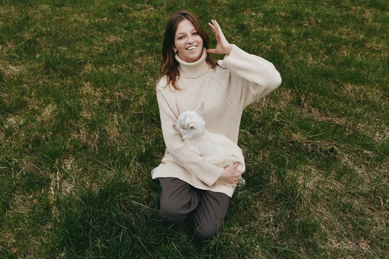 Cheerful woman outdoors holding a goat kid in a sunlit field, cozy sweater, relaxed smile, gentle breeze, natural colors, lush grass, rural lifestyle moment
