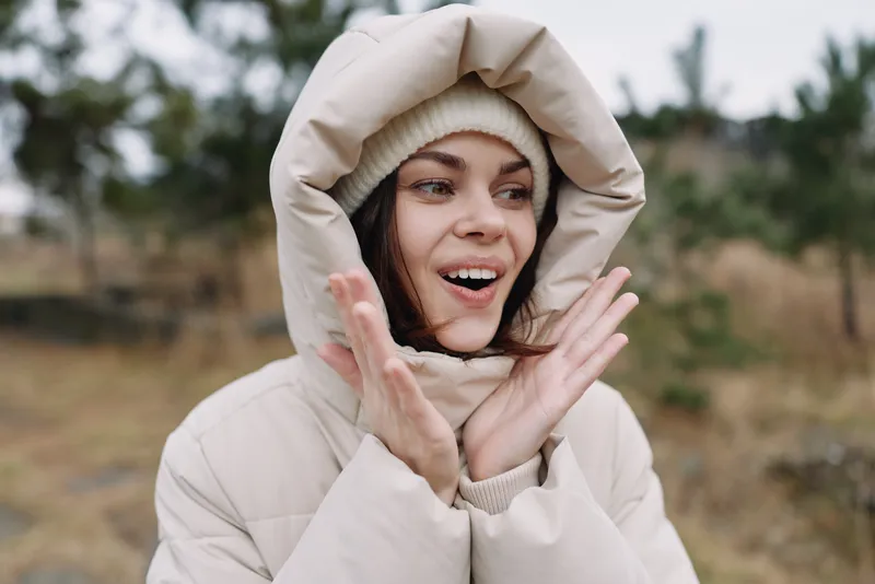 Woman in a warm hooded coat outdoors with a surprised expression, enjoying chilly weather in a natural landscape, candid and joyful moment.