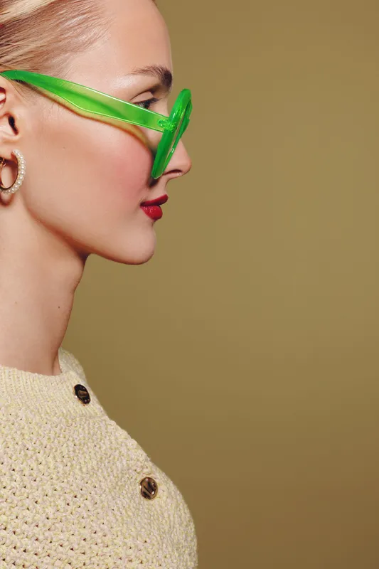 Profile of a stylish woman wearing green transparent glasses and a knitted beige sweater with buttons. Minimalist studio portrait on a plain olive background.