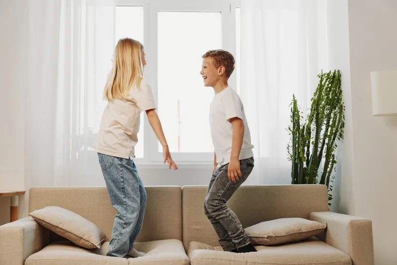 Two happy children jumping on a beige sofa in bright living room with natural light. Concept of childhood fun, family moments, and indoor playtime.