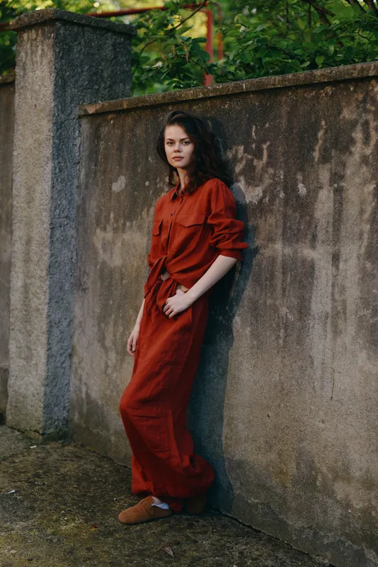 Casual woman in a rust red jumpsuit stands against a weathered concrete wall outdoors, creating a relaxed lifestyle portrait with natural light, moody urban scenery and a calm, thoughtful mood