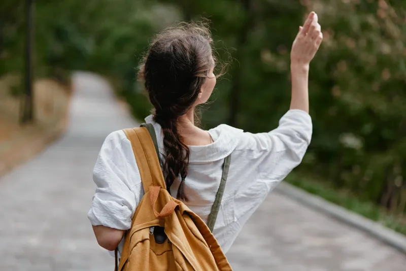 A young woman waving goodbye while walking on a tree lined path, showcasing casual, stylish outdoor attire and a sense of optimism