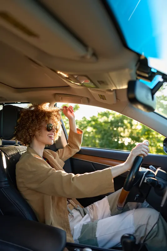 Curly haired woman driving a car with a retro film color palette, casual chic style, sunny daytime, candid lifestyle vibe, focus on mobility, independence, and warm mood.