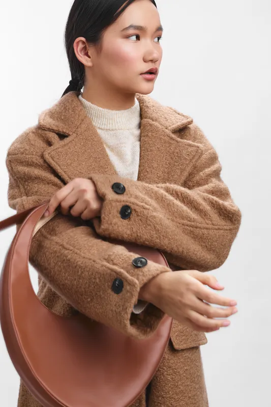 asian woman wearing a tan wool coat holds a curved handbag, standing in a bright studio. she looks confident and poised, presenting a modern chic aesthetic.