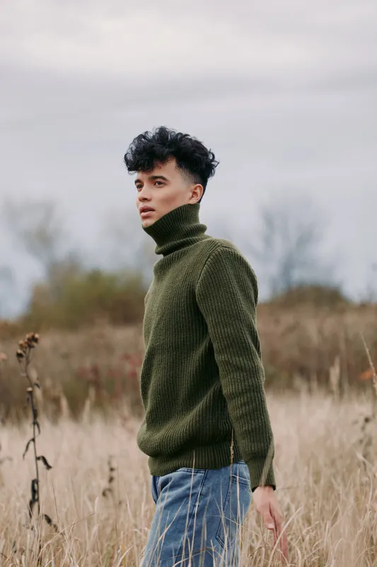 Young man in a textured green sweater stands in a windswept field, conveying a natural, honest portrait of everyday style and quiet confidence in a candid outdoor moment.