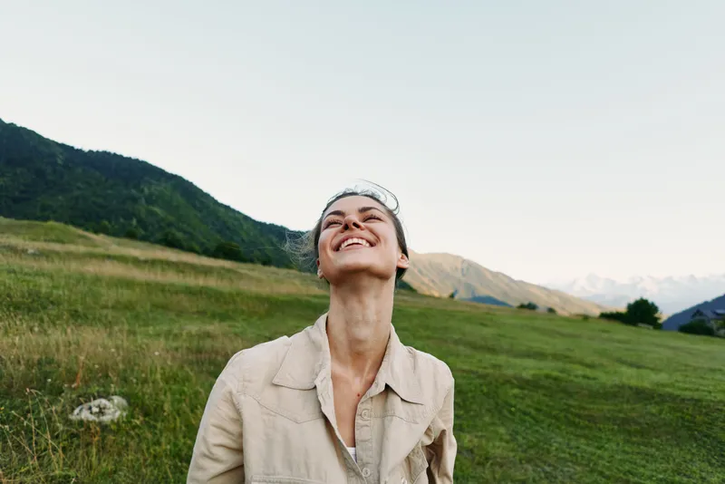 Cheerful woman outdoors, smiling up at the sky in a sunlit meadow, relaxed posture, natural colors, wide landscape, mountains in distance, lifestyle moment