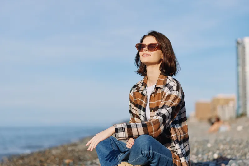Woman on a sunny beach wearing sunglasses and a plaid shirt, seated in a relaxed pose for a casual outdoor portrait by the sea and horizon.