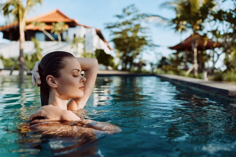 Relaxed woman floats in a pool with eyes closed, arms resting on her shoulders, enjoying calm water and warm sun, tropical resort surroundings, serene mood, leisure swim, vacation atmosphere