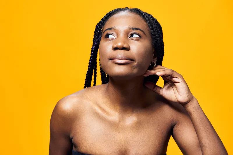 Young Black woman with braided hair expressing curiosity against a vibrant yellow background, showcasing natural beauty and confidence in a minimalist style