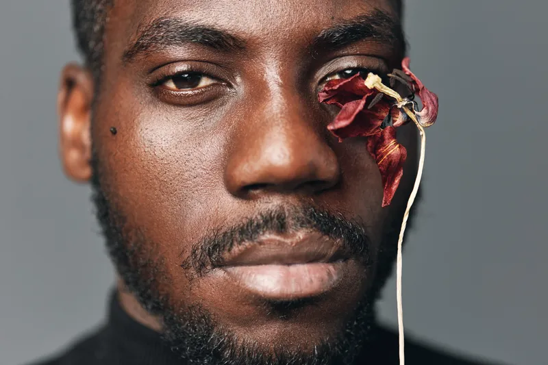 African man with dark skin and beard in creative conceptual style closeup portrait with dried red flower covering one eye on gray background for visual art and expression.
