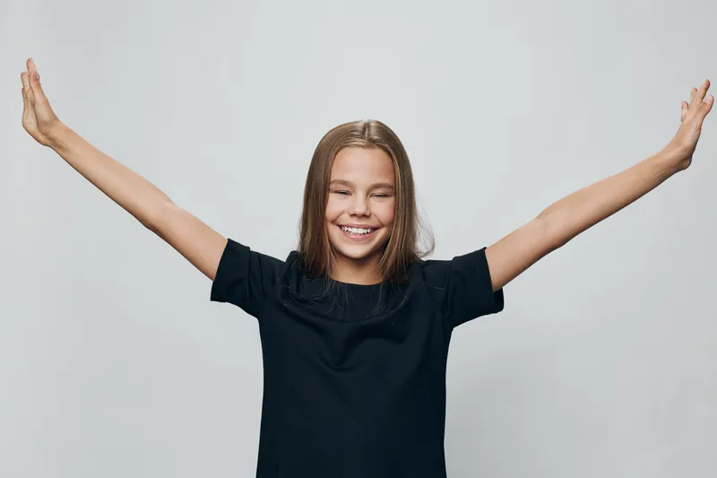 Girl with long hair smiling and stretching arms wide in black shirt on white background, happiness and joy expression with closed eyes and relaxed posture.