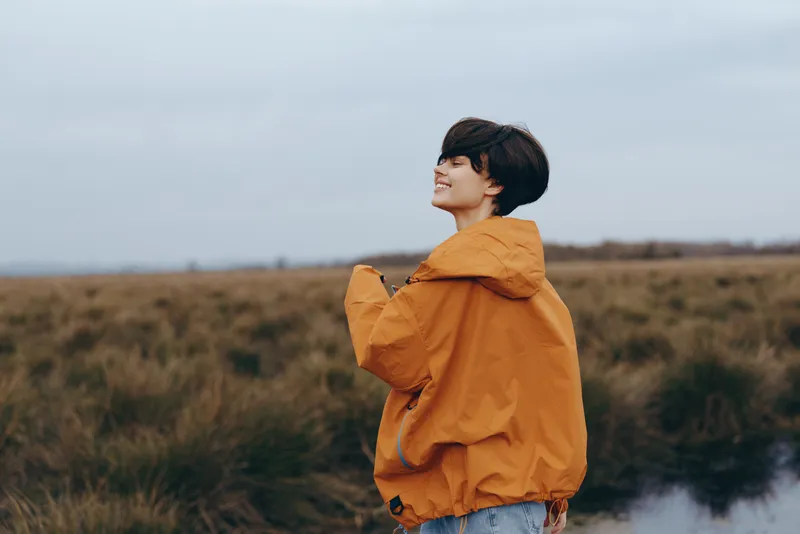 Woman lifestyle portrait outdoors by a river, orange hooded jacket, denim jeans, side view smiling, cloudy sky, calm travel mood, riverbank at dusk with misty water and open fields