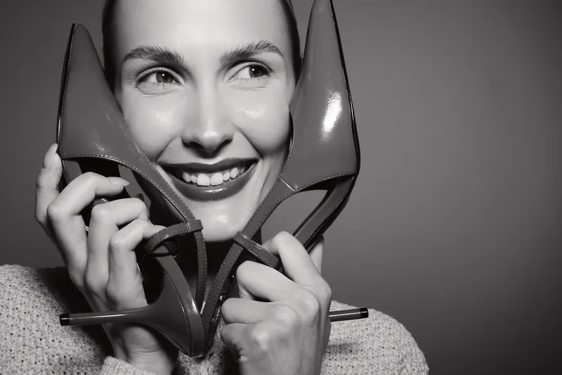 Smiling Caucasian woman with light skin holds two high heel shoes near her face in black and white photo. Concept of fashion, happiness, and elegance.