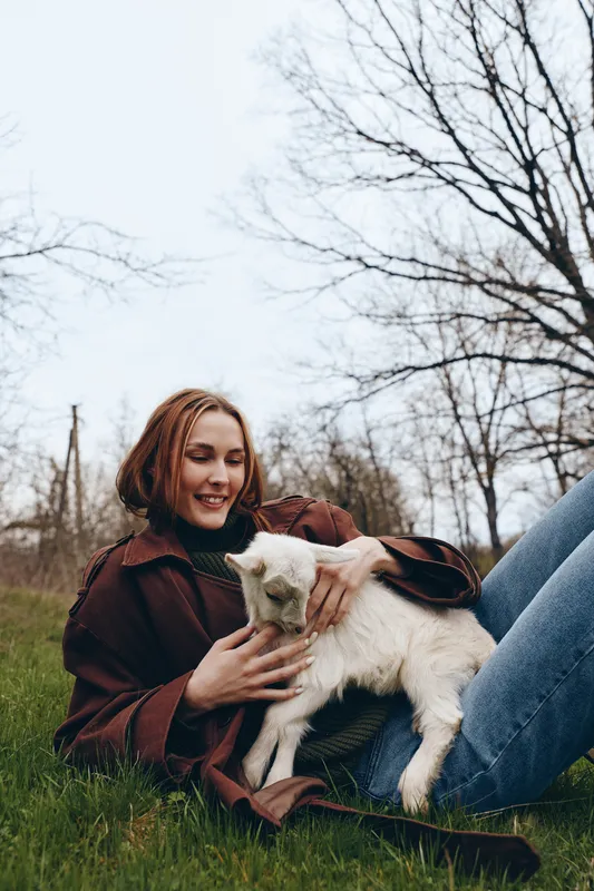 A young woman with short brown hair smiles gently while sitting on grass and holding a small white baby goat in a bare tree outdoor setting during early spring.