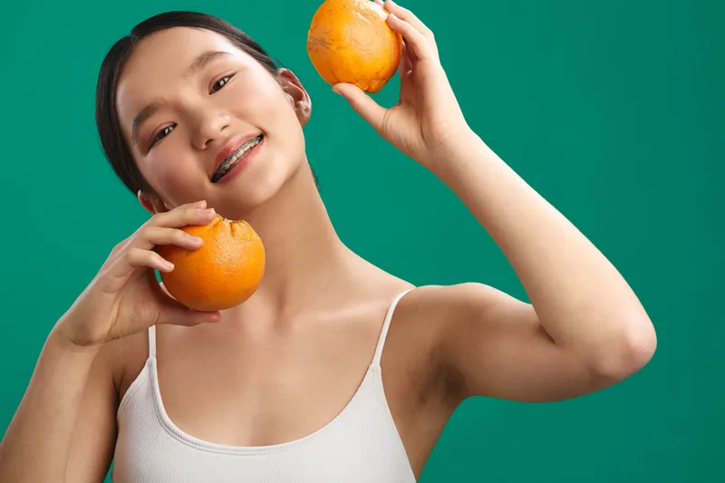 asian woman smiles broadly while holding bright oranges against a vivid green background, delivering a fresh, cheerful vibe and a healthy, energetic mood.