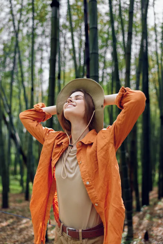 Woman in hat with a smile breathes fresh air in bamboo forest, nature escape with relaxed pose, closed eyes and orange jacket enjoying outdoor serenity and sunlight.