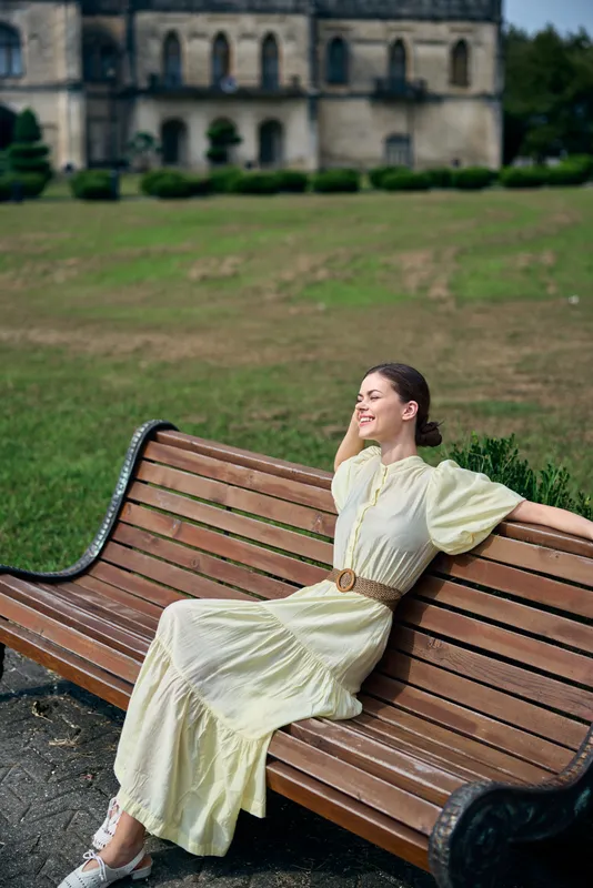 A woman in a pale yellow dress sits on a wooden bench in a sunny park, with a distant historic castle, exuding elegance, poise, and relaxed outdoor charm.