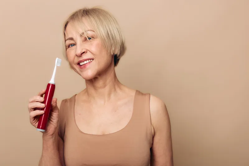 Smiling mature woman with short blonde hair holds electric toothbrush against beige background. Concept of dental care, oral hygiene, healthy lifestyle, and self-care for older adults.