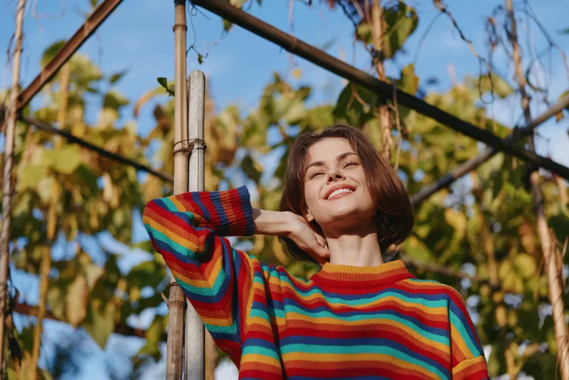 Woman smiling in rainbow sweater outdoors among autumn vines and nature, portrait of happy young female enjoying sunlight, casual colorful knit and relaxed pose.