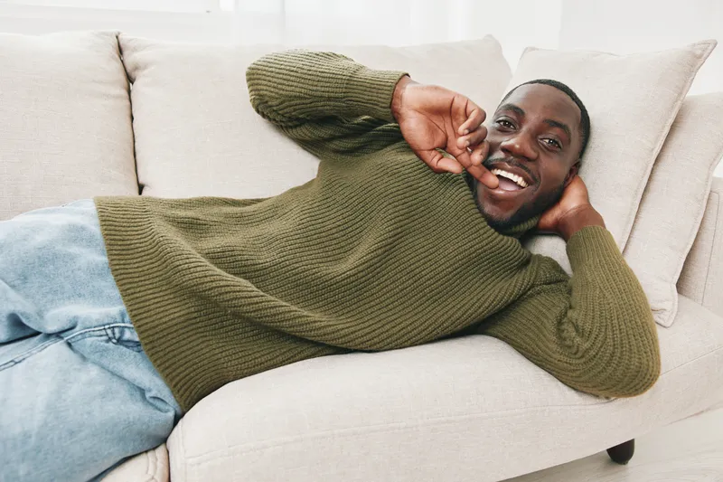 Happy young African man relaxing on a cozy sofa, wearing a knitted sweater and casual jeans, exuding warmth and comfort in a bright, inviting living space