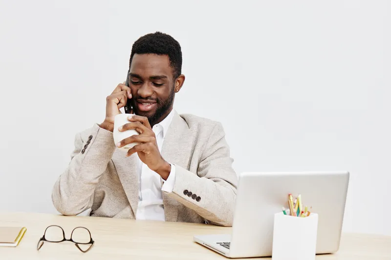 Smiling man talking on mobile phone during work at desk in modern office, dressed in beige blazer, positive mood, professional atmosphere, using technology, natural light. People lifestyle concept, co