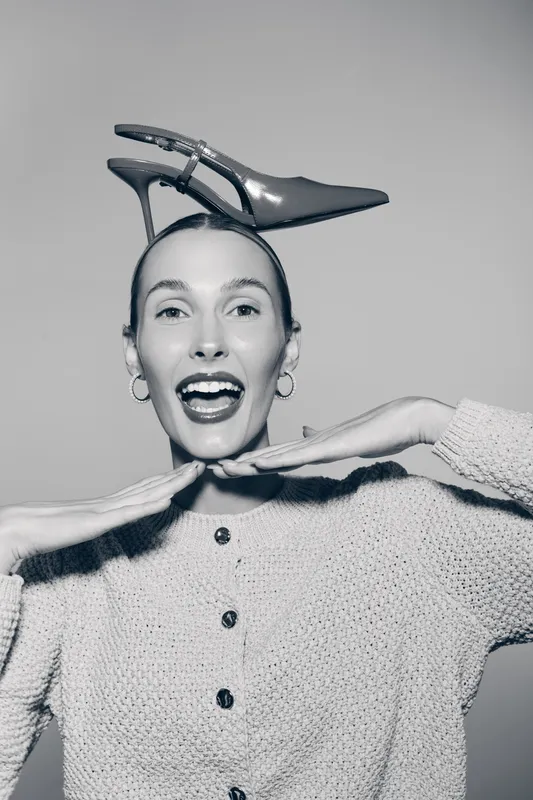 Happy young white woman with light skin and high cheekbones smiles widely wearing a shoe on her head. Concept for fun, fashion, and playful mood in studio.
