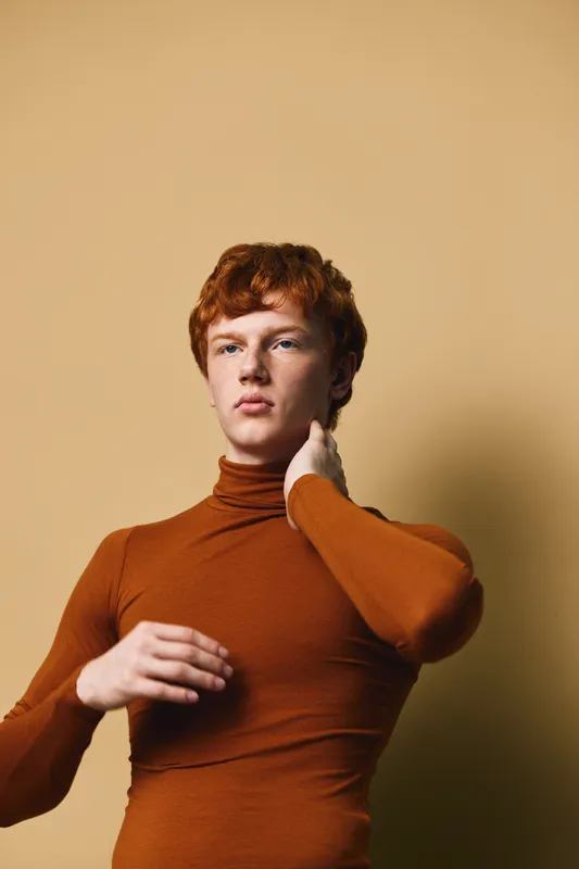 Young man with red hair wearing brown turtleneck posing against beige background, looking thoughtful with one hand touching neck, studio portrait in soft natural light.
