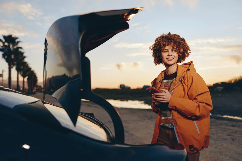 Smiling woman with curly hair stands outdoors beside a car at sunset, holding a jacket. Lifestyle travel moment on a road, warm golden light at dusk.