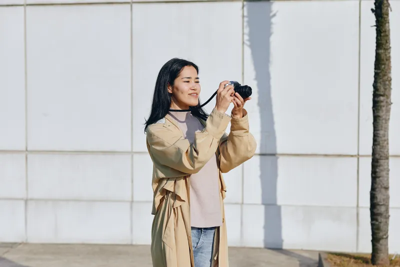Woman posing with camera outdoors, capturing moments in daylight, wearing a long beige coat and soft sweater, casual street photography scene, lifestyle and travel vibe
