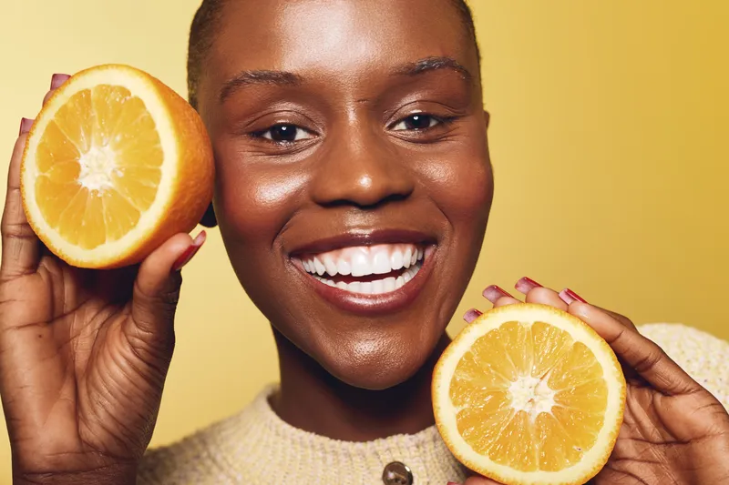 Happy smiling African woman with dark skin holds two halves of fresh orange fruit close to her face on yellow background. Concept for healthy lifestyle, vitamin C, and natural beauty.