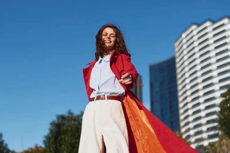 Young woman with curly hair smiles confidently while wearing a red coat and white pants outdoors in an urban setting under clear blue sky.