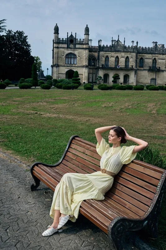 woman on bench in park near historic estate and architecture, wearing flowing dress and relaxing with hands behind head, authenticity inclusivity, leisure scene with calm grounds