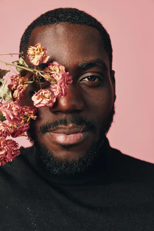 African man with dark skin and beard posing with dried pink flowers covering one eye in creative conceptual visual style. Closeup portrait with soft pink background and black turtleneck clothing.