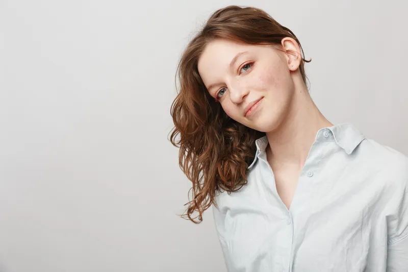 Young woman with curly brown hair smiles gently wearing a light blue shirt against a plain light background. Concept of natural beauty, casual style, and confident female portrait.