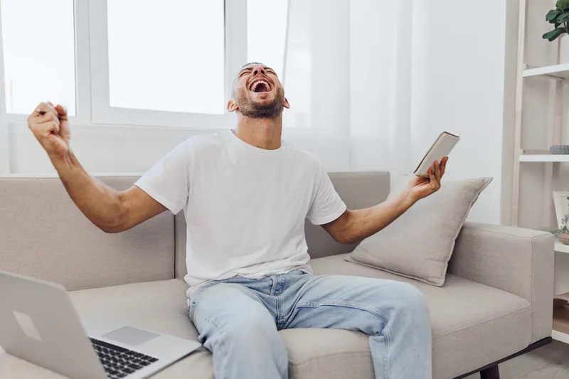 Joyful man celebrating a successful project while juggling work-life balance, showcasing authentic emotions and soft masculinity in a cozy home setting.