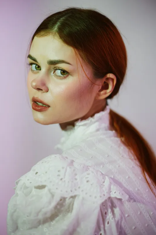 A captivating young woman with long brown hair in a vintage white dress against a soft purple background, showcasing a contemplative expression and elegant style