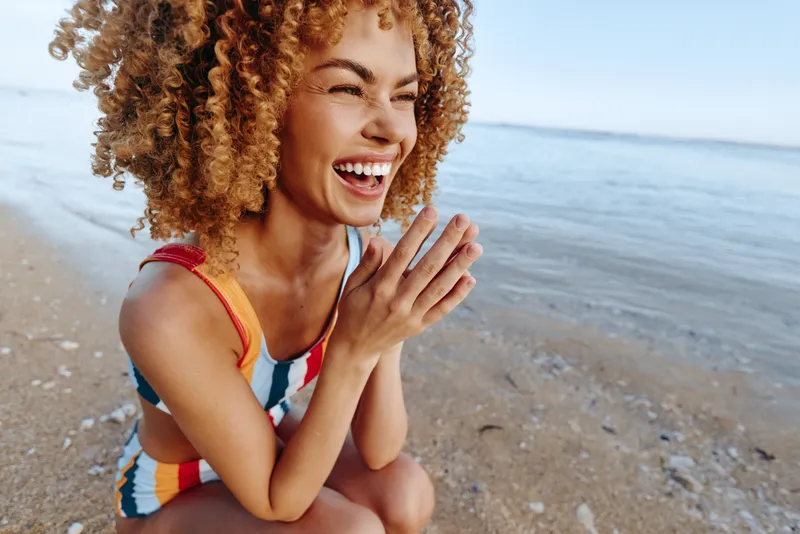 Smiling woman with curly hair on a beach by the ocean, hands near face, laughing in sunlight, casual summer travel mood, shallow depth of field, natural skin tones relaxed pose.