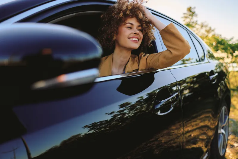 Cheerful woman in a car leaning out the window, sunlit outdoors moment with a relaxed pose, vintage lifestyle vibe, curly hair and casual fashion against greenery