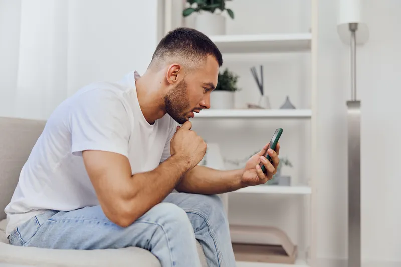 Thoughtful man engaged with smartphone, exploring digital content in a minimalist living room filled with greenery and modern decor. Ideal for tech-related themes.