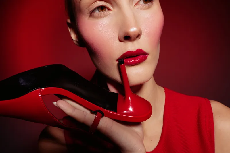 Close-up of a woman with red lipstick holding a red high heel shoe near her face against a red background. Concept of fashion, beauty, style, and elegance in studio lighting.