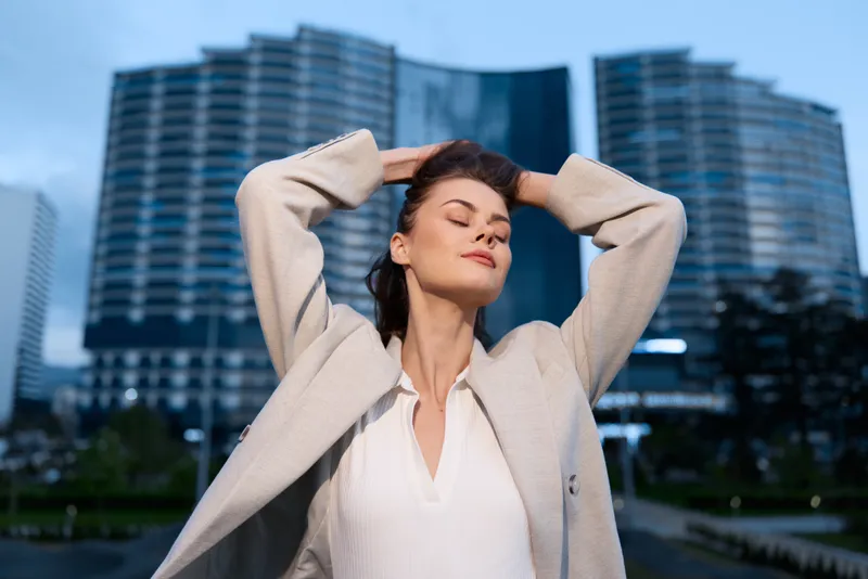 Confident woman with closed eyes in beige coat feeling relaxed outdoors in urban business district during daylight