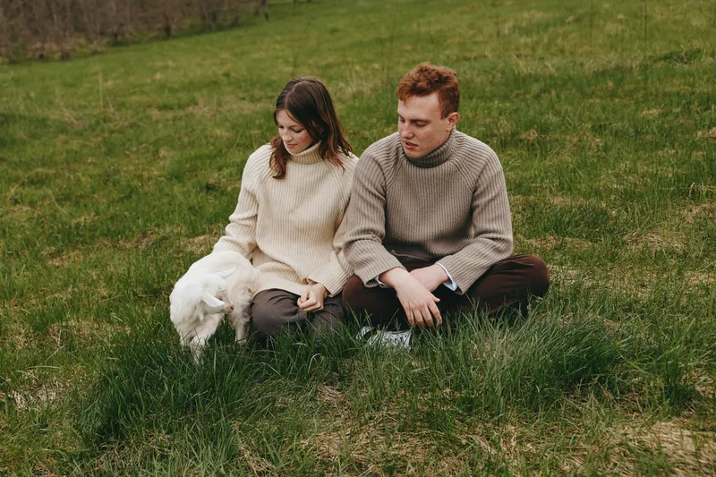 Couple outdoors with a goat kid on the grass, wearing sweaters, enjoying a cozy lifestyle and peaceful moment in nature.
