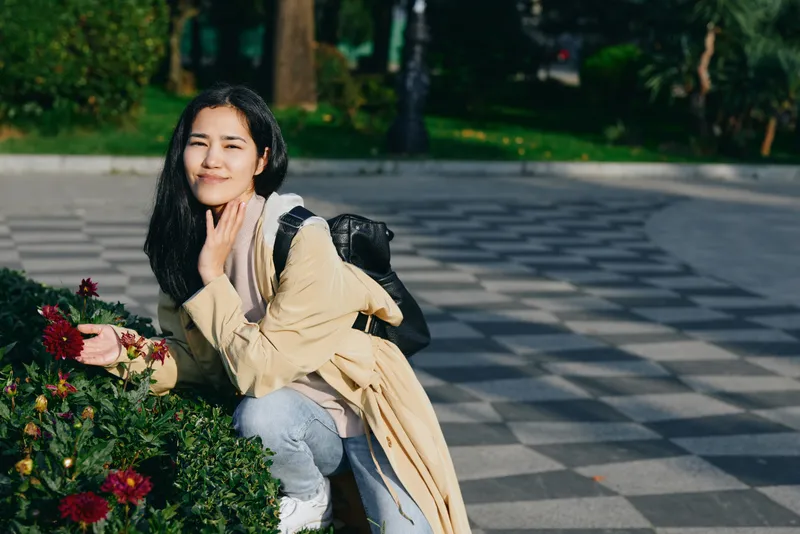 Woman crouching beside blooming flowers in a park, enjoying a sunny day, casual fashion, backpack ready for a stroll, candid moment of calm and beauty.