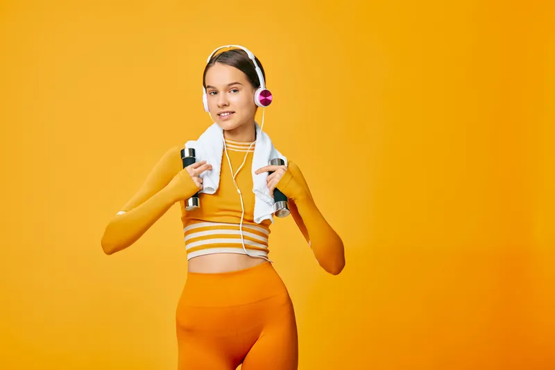 Fitness woman in bright orange athletic set, wearing headphones, with a towel around neck and two bottles, standing against a bold orange backdrop, ready for workout, energy, and motivation.