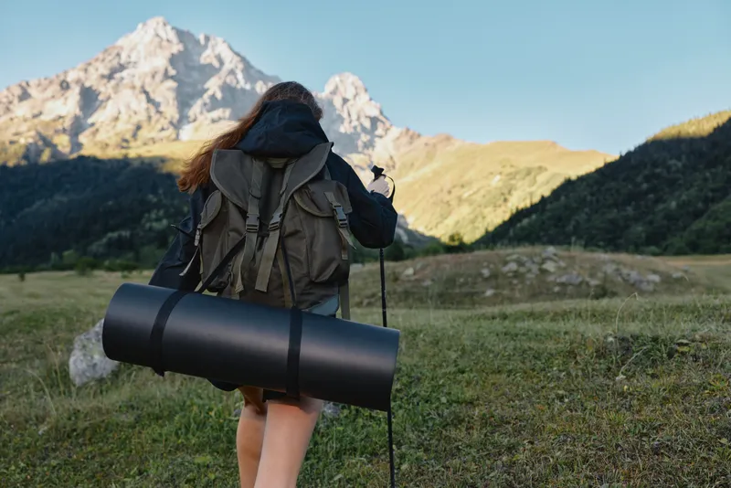 A lone hiker with a backpack and trekking pole walks toward a rugged mountain peak on a clear day, showcasing adventure, exploration, and outdoor dedication.