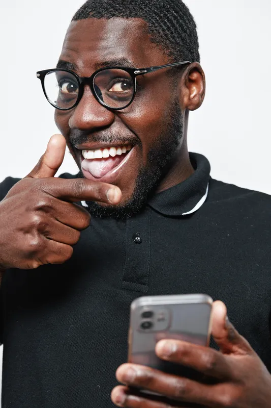 African American man with dark skin and glasses smiling and pointing finger while holding smartphone in black polo shirt on white background. Positive expression and casual style.
