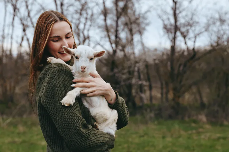 Cheerful woman outdoors, embracing a young goat kid, warm moment, earthy tones, gentle breeze, natural backdrop of fields and trees, close connection with animals, rustic lifestyle