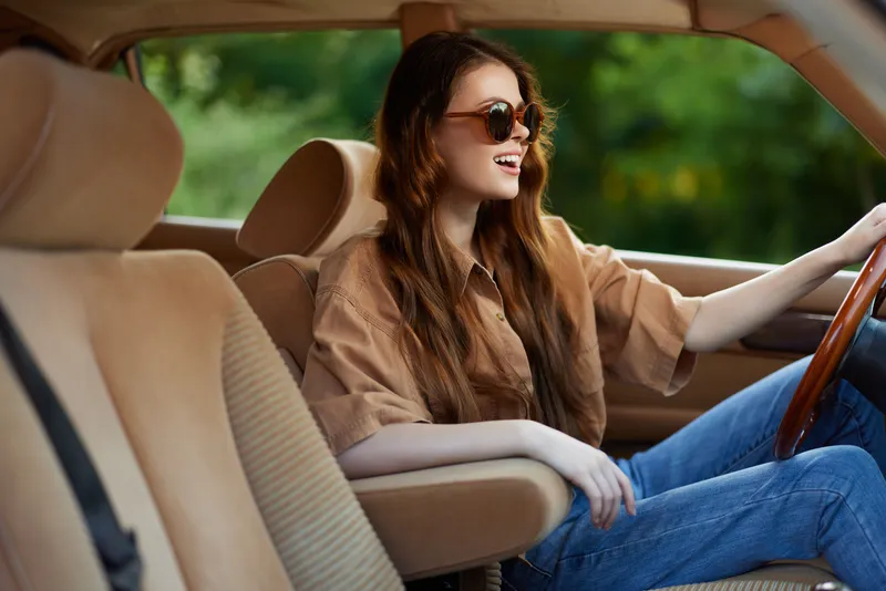 Young woman with long hair in stylish sunglasses smiling while driving a vintage car on a sunny day, conveying a sense of freedom and joy.