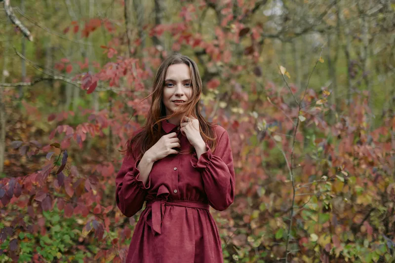Young woman in a deep red dress stands in an autumn forest holding a leaf close, an authentic portrait capturing genuine emotion with natural light and realistic seasonal tones.